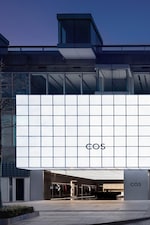 Placeholder for: Modern building with a large, illuminated grid facade and "COS" logo. Store entrance below with visible interior displaying clothing.