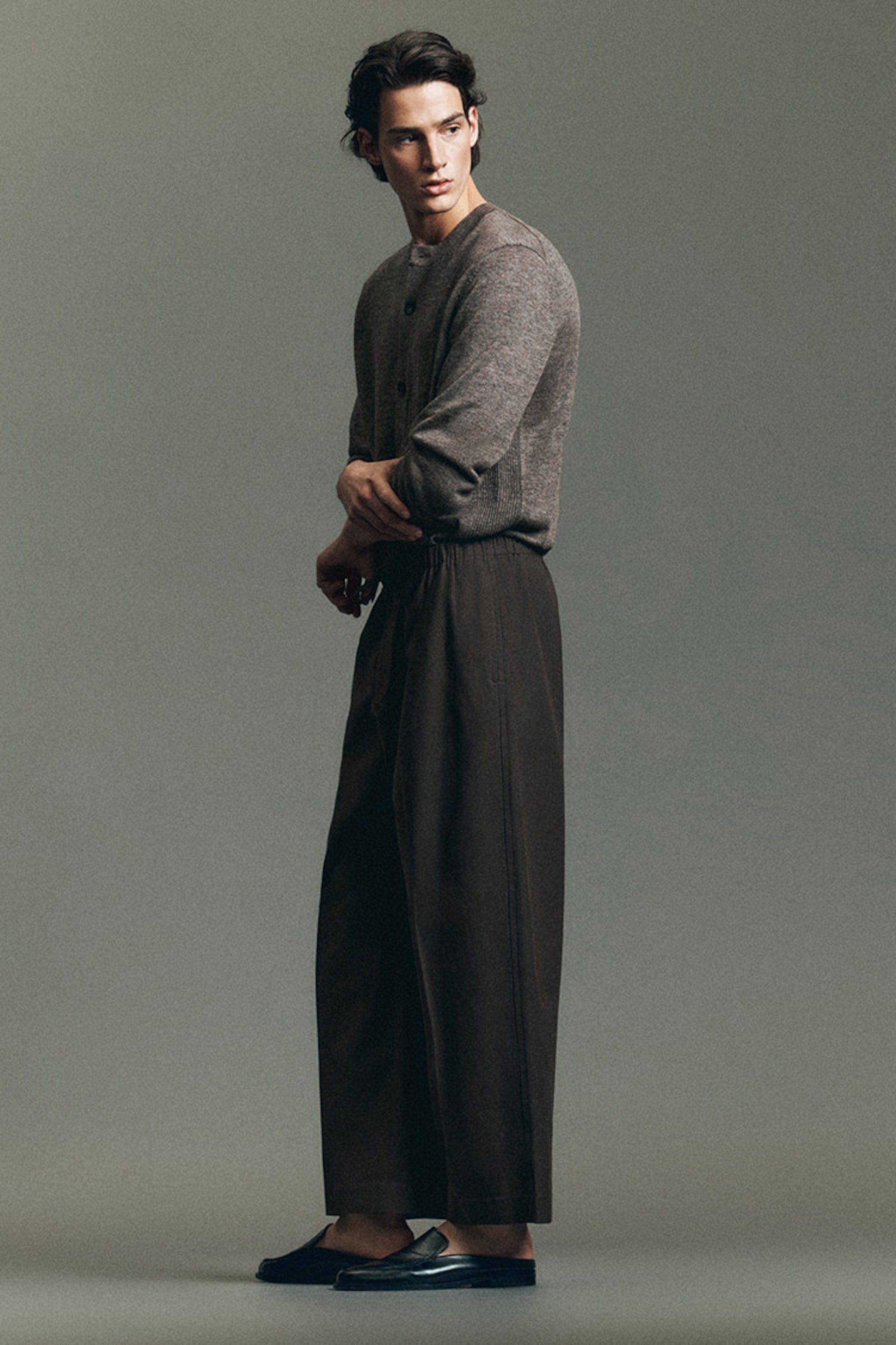 Man in a gray sweater and brown wide-leg pants poses in a studio setting, looking to the side.
