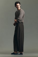 Placeholder for: Man in a gray sweater and brown wide-leg pants poses in a studio setting, looking to the side.