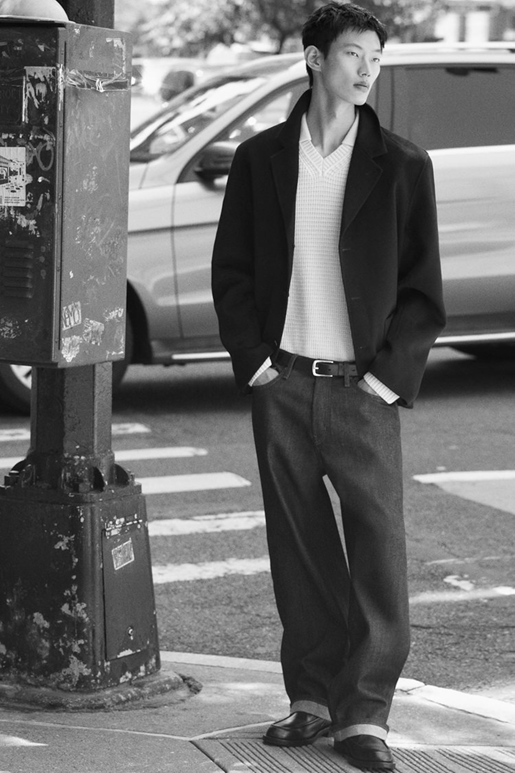 A man in a black jacket and white shirt stands casually on a city street corner, hands in pockets, looking sideways.