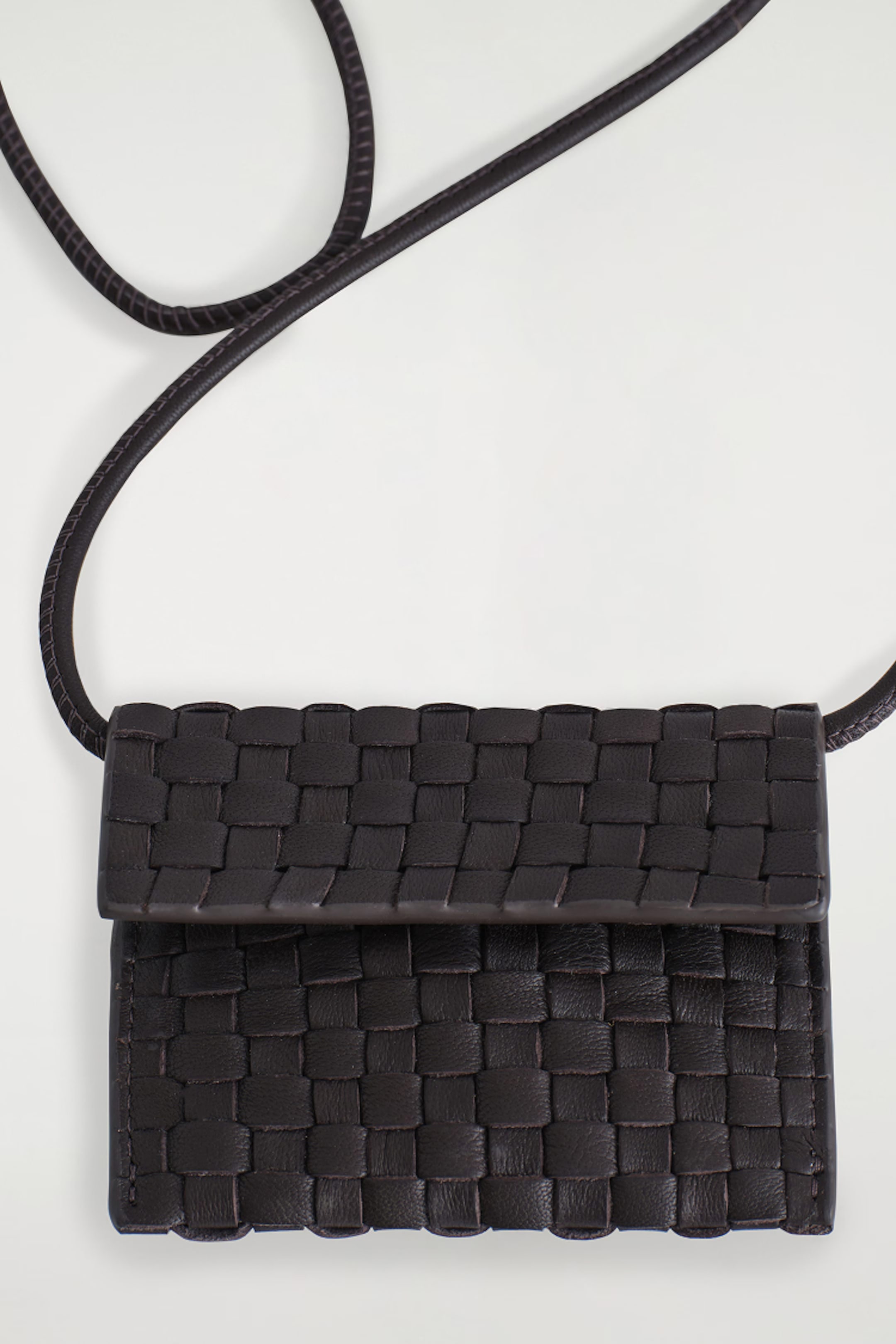 A black woven leather crossbody bag with a flap closure and adjustable strap, set on a light gray background.