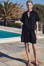Placeholder for: A person in a black dress stands by a pool, with palm trees in the background on a sunny day.