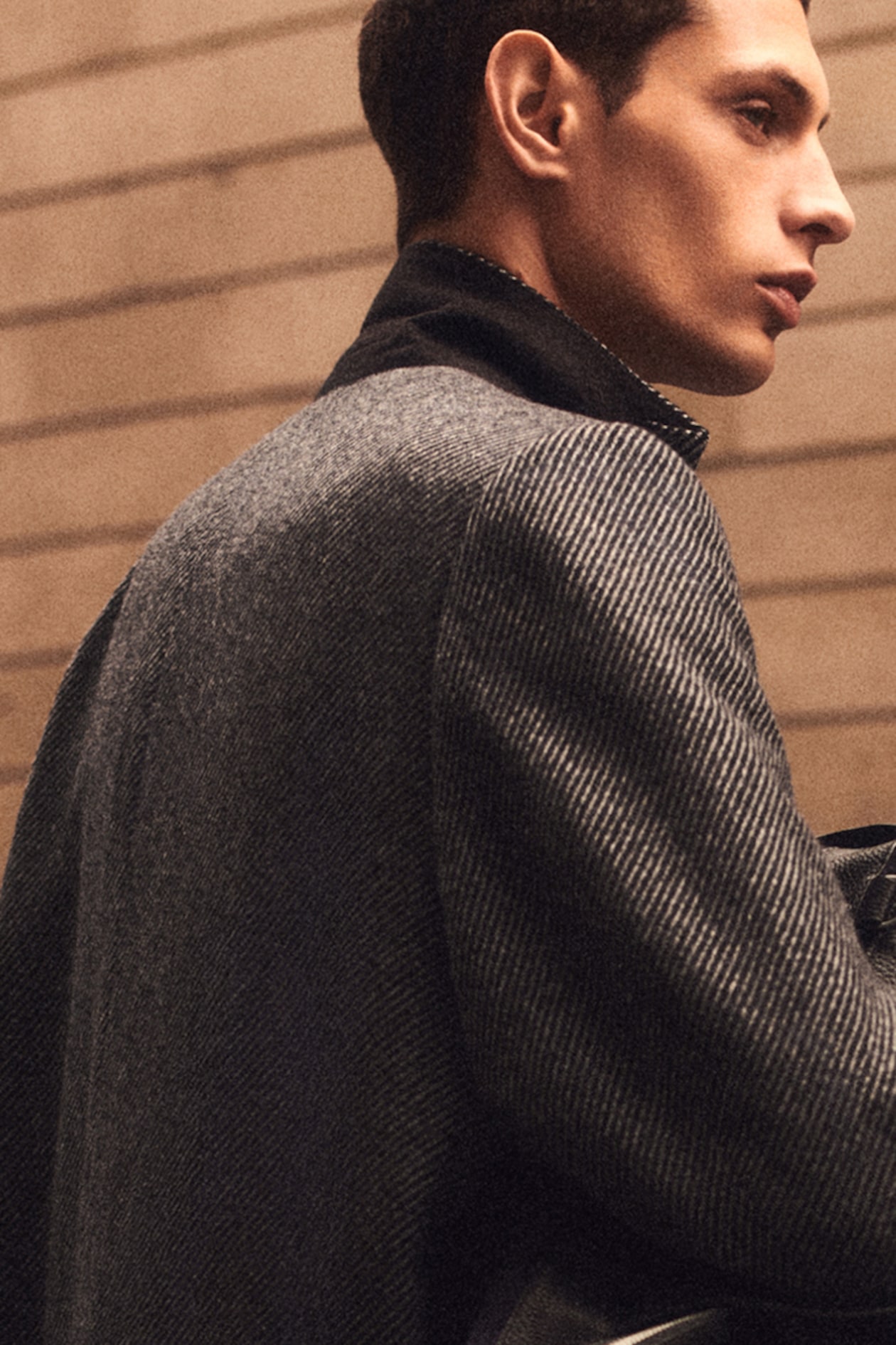 Man in a stylish gray herringbone coat with a black collar, standing sideways against a blurred, neutral-toned background.