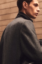 Placeholder for: Man in a stylish gray herringbone coat with a black collar, standing sideways against a blurred, neutral-toned background.