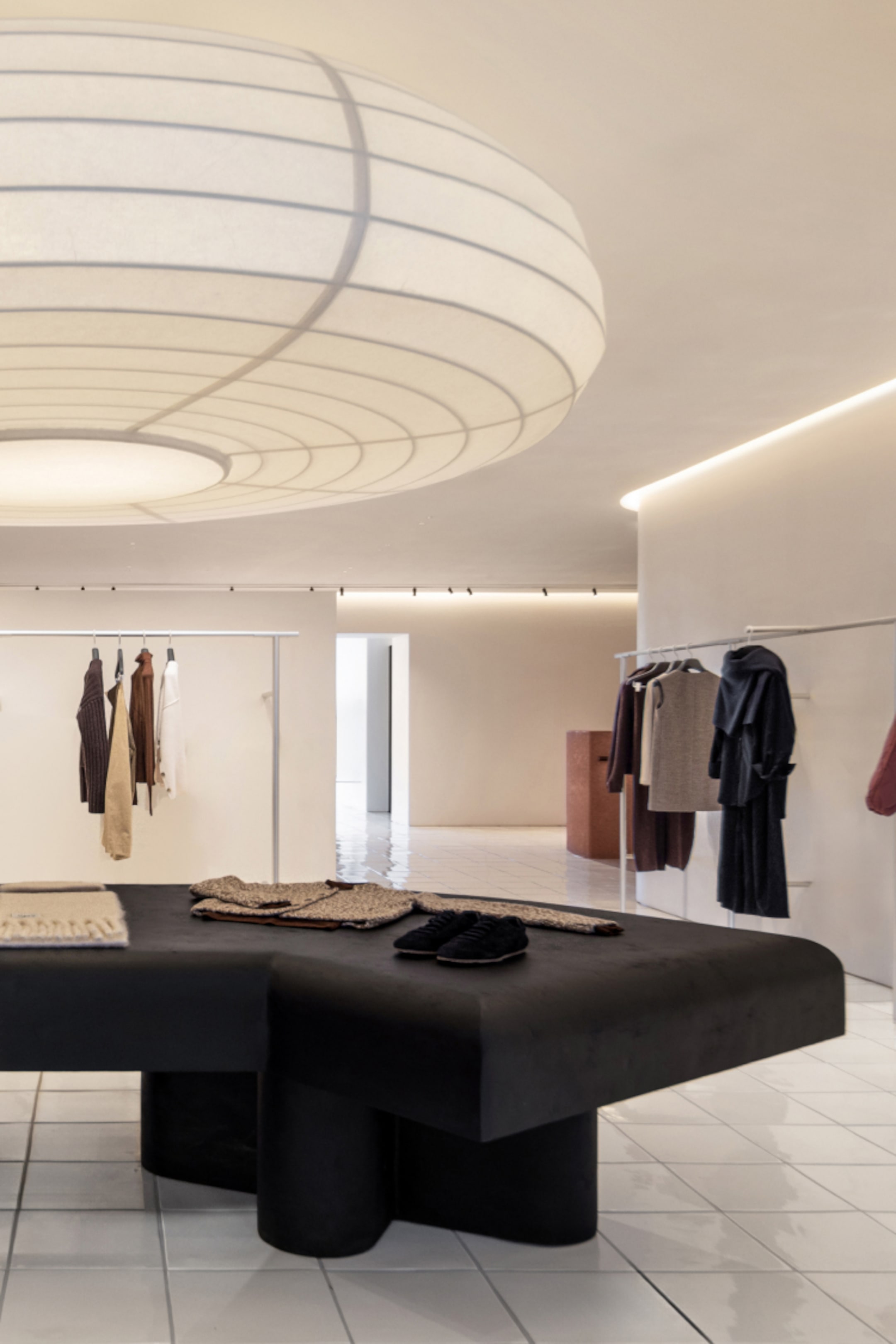 Minimalist boutique interior with hanging clothes, a large black table displaying items, a white tiled floor, and a large round ceiling light.