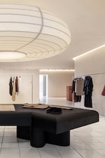 Placeholder for: Minimalist boutique interior with hanging clothes, a large black table displaying items, a white tiled floor, and a large round ceiling light.