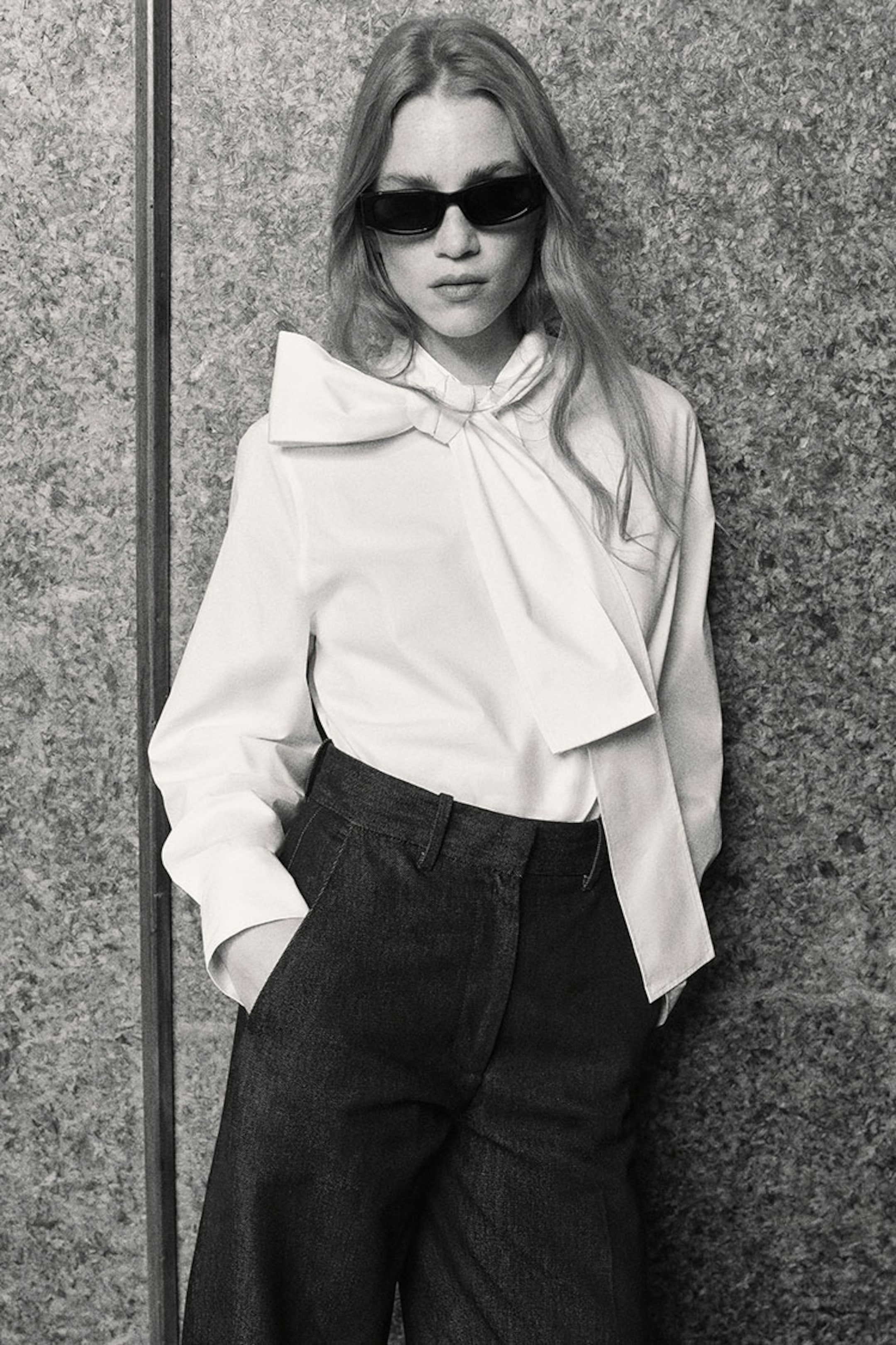 Woman in sunglasses and a white blouse with a large bow, wearing dark pants, stands against a textured wall.