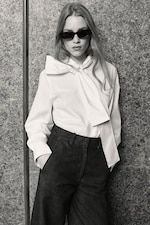 Placeholder for: Woman in sunglasses and a white blouse with a large bow, wearing dark pants, stands against a textured wall.