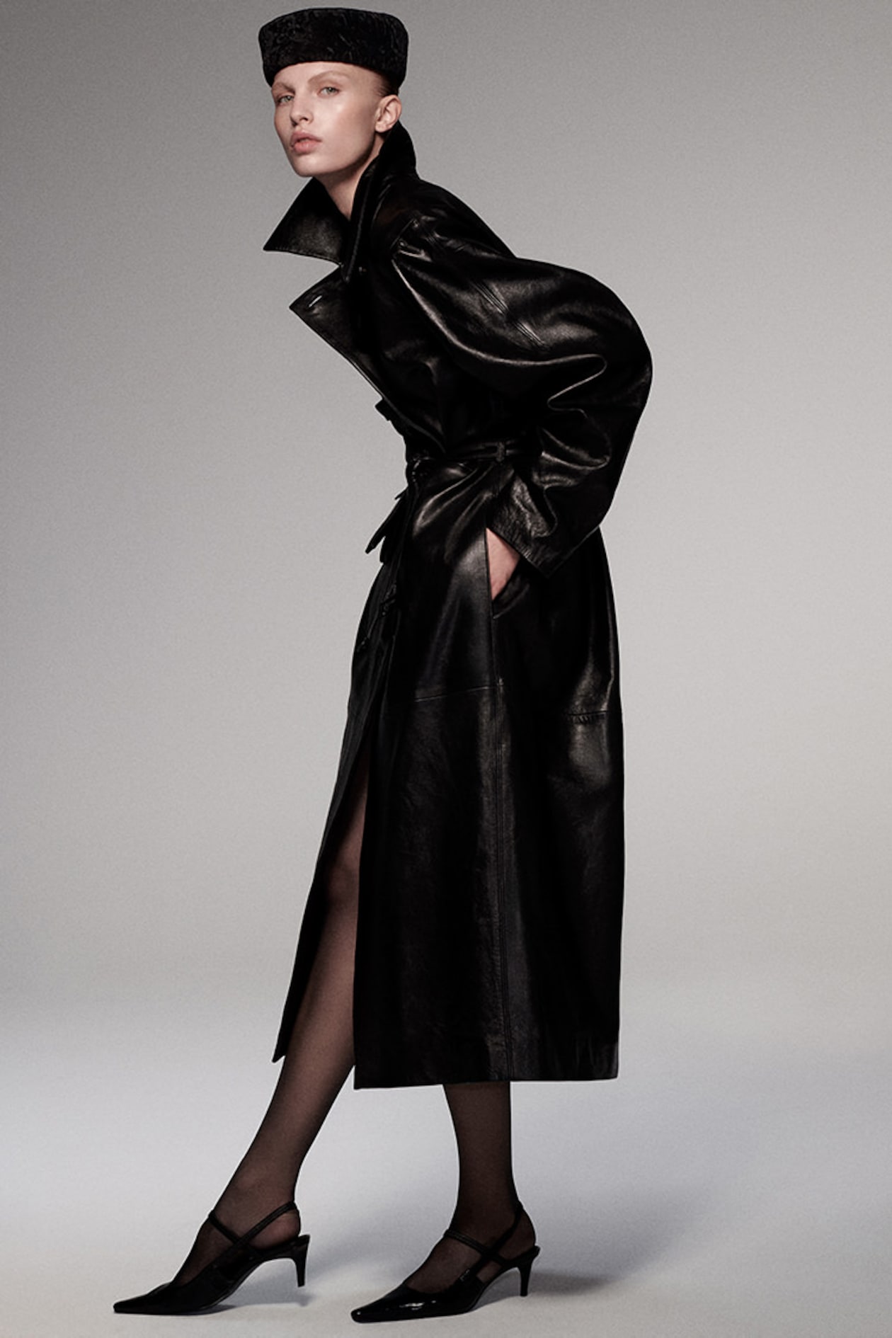 A person wearing a long black leather coat, a dark hat, and black heels stands in a fashion pose against a plain background.