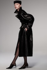 Placeholder for: A person wearing a long black leather coat, a dark hat, and black heels stands in a fashion pose against a plain background.