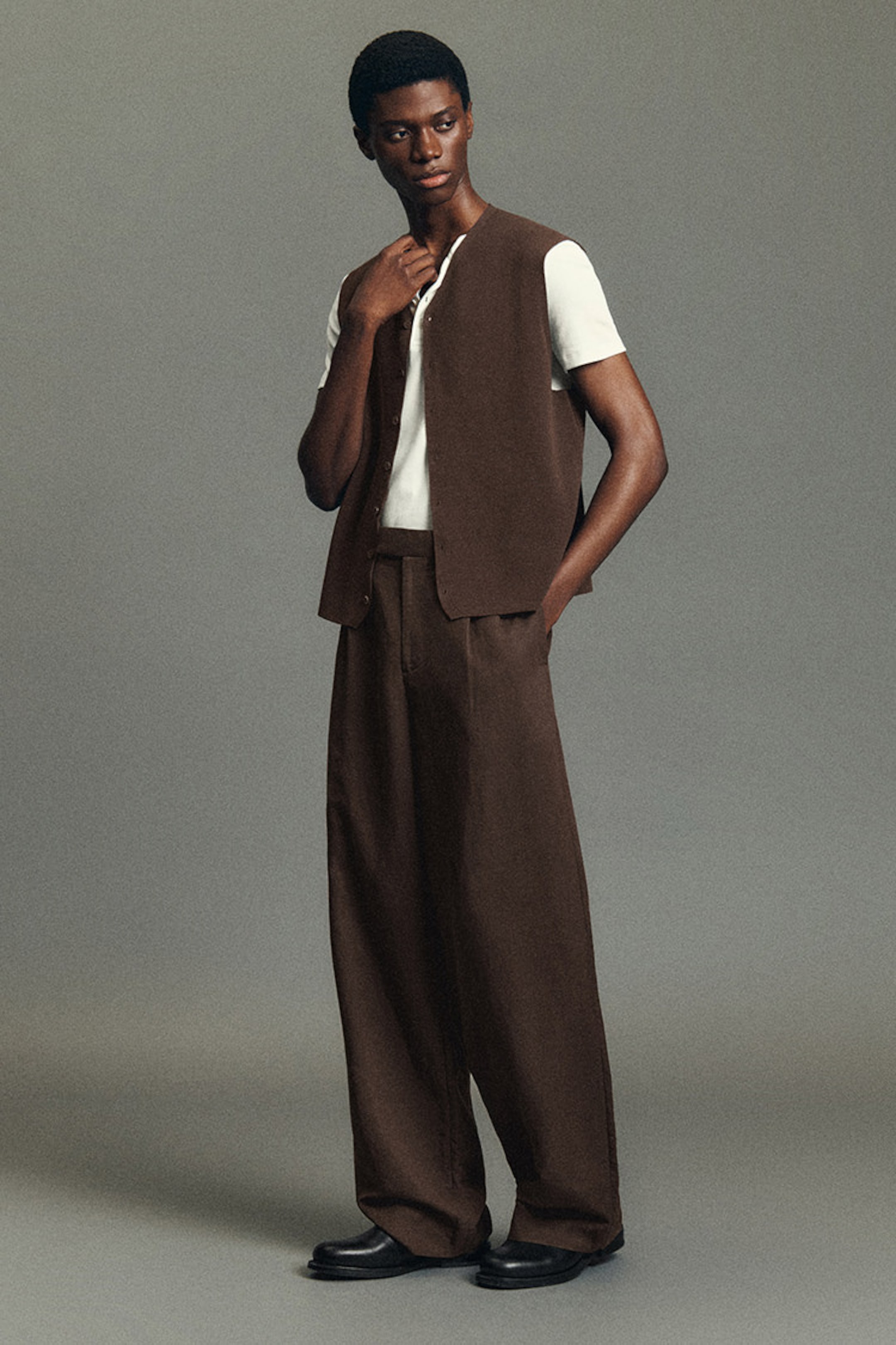 Person in a brown vest and wide-legged pants, wearing a white shirt, stands thoughtfully against a neutral backdrop.