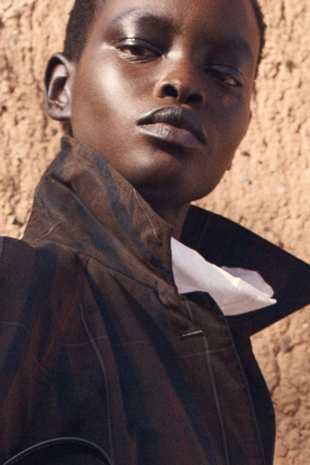 Person in a brown, striped jacket with a white shirt beneath, standing against a textured, sandy wall, looking upward confidently.