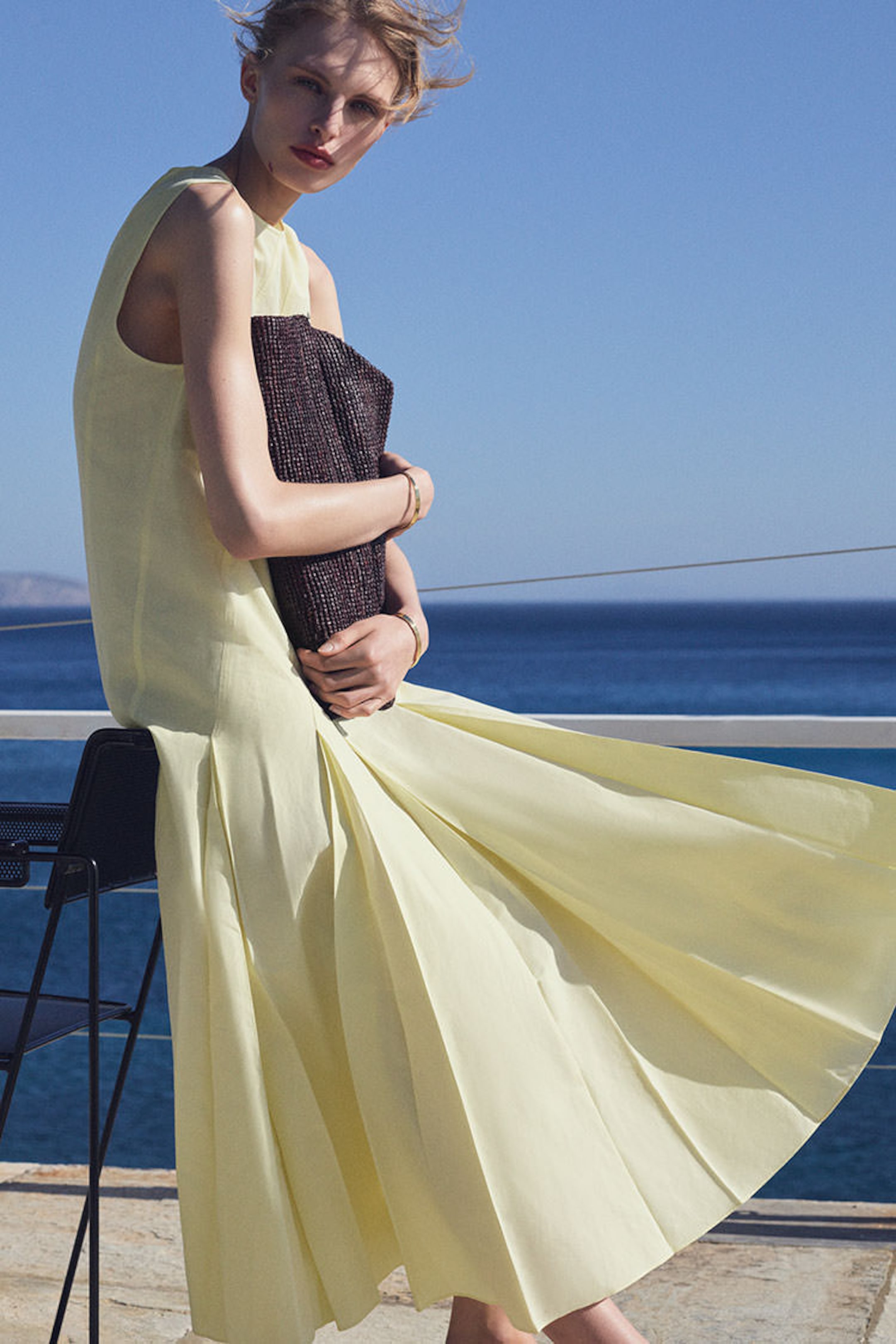 Person in a flowing yellow dress holding a clutch, seated outdoors near a railing with a sea view.