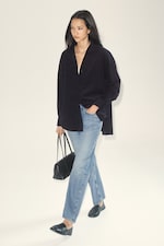 Placeholder for: Person in a black shirt and blue jeans walking while carrying a black handbag, against a plain background.