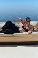 Placeholder for: Person lounging on a beige sofa by the sea, wearing sunglasses and a black outfit. The ocean and sky form a serene backdrop.