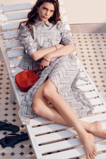 Placeholder for: Woman in a patterned dress lounges on a white chaise lounge, holding a red bag, with sandals nearby on a tiled floor.