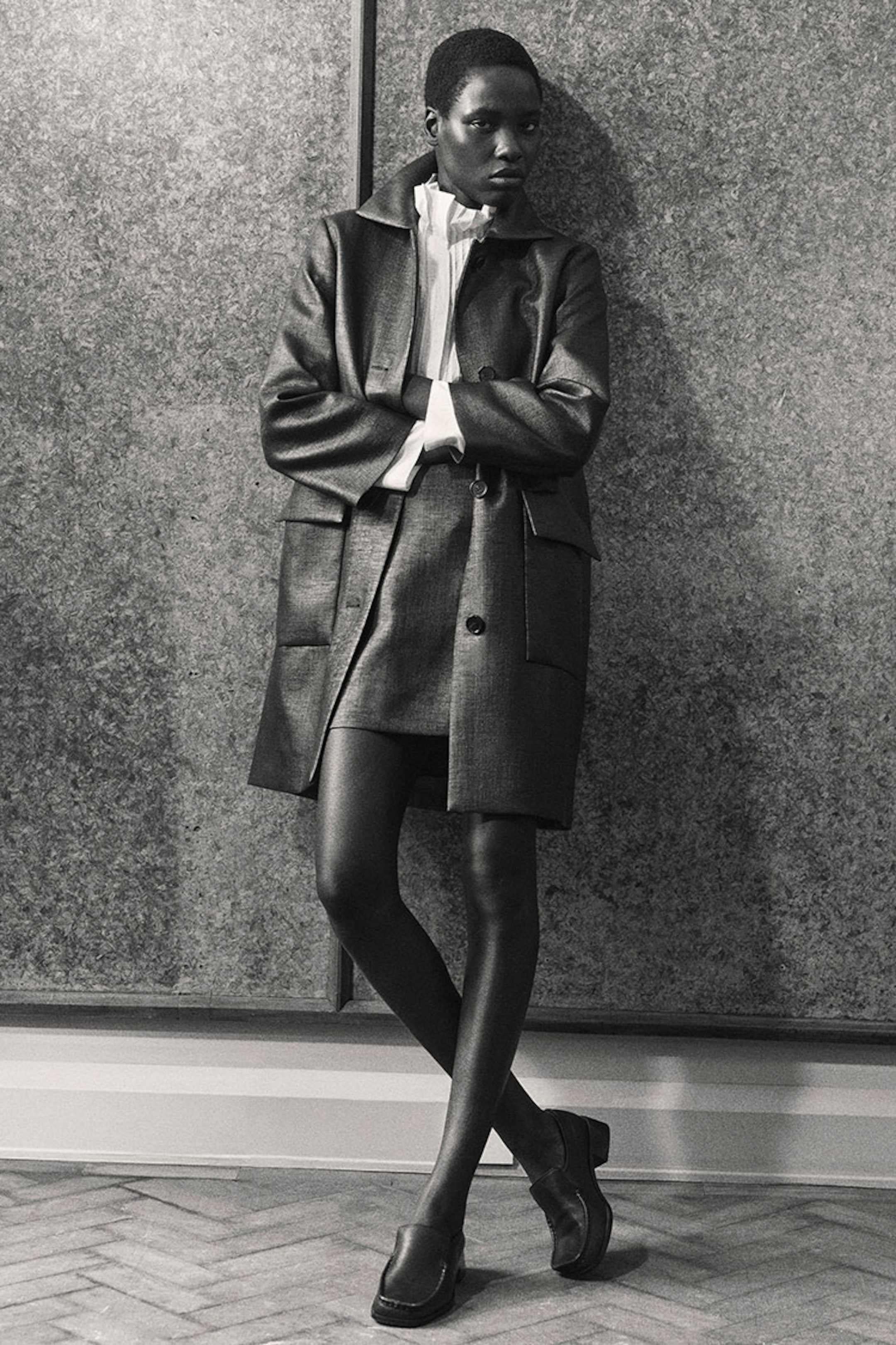 Person in a stylish, dark coat and skirt ensemble, leaning against a textured wall, with arms crossed and wearing loafers.