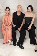 Placeholder for: Three people posing in stylish outfits; two women in elegant dresses and a man in a black suit, seated against a marble backdrop.
