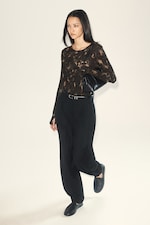 Placeholder for: Person wearing a dark, patterned top, black pants, and shoes, holding a black bag under their arm, standing against a plain background.