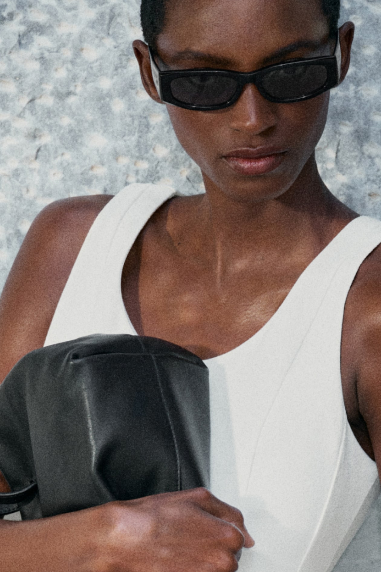Person in sunglasses wearing a white top, holding a black leather bag. The background is a textured, light-colored surface.