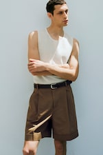 Placeholder for: Man in a sleeveless white shirt and brown shorts with a black belt, standing against a light-colored wall, arms crossed.