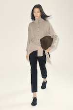 Placeholder for: Woman in a beige sweater and black pants walks confidently, holding a dark brown clutch. She has long black hair and wears black shoes.