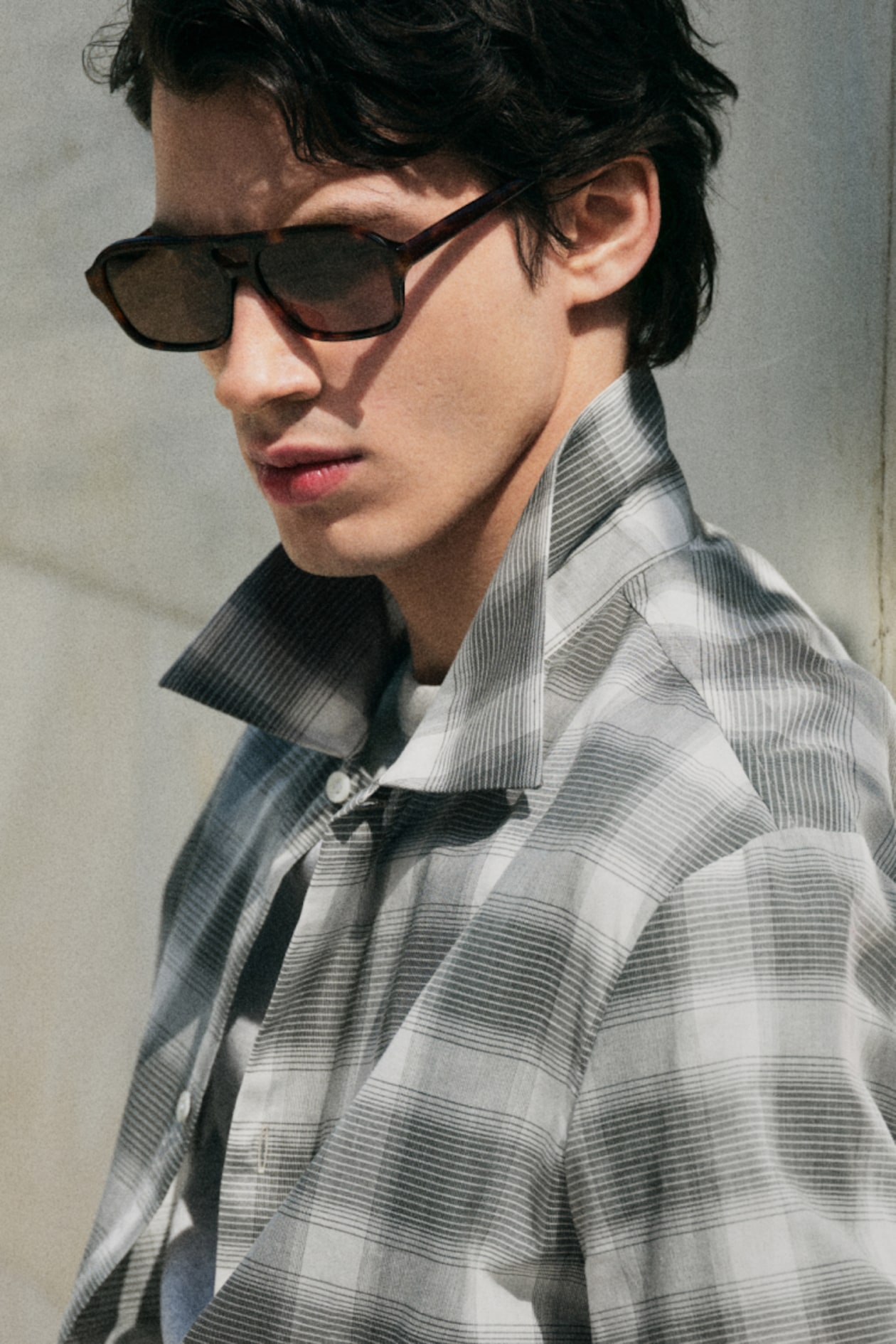 Man in sunglasses wearing a plaid shirt with a raised collar, gazing sideways.
