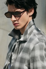 Placeholder for: Man in sunglasses wearing a plaid shirt with a raised collar, gazing sideways.
