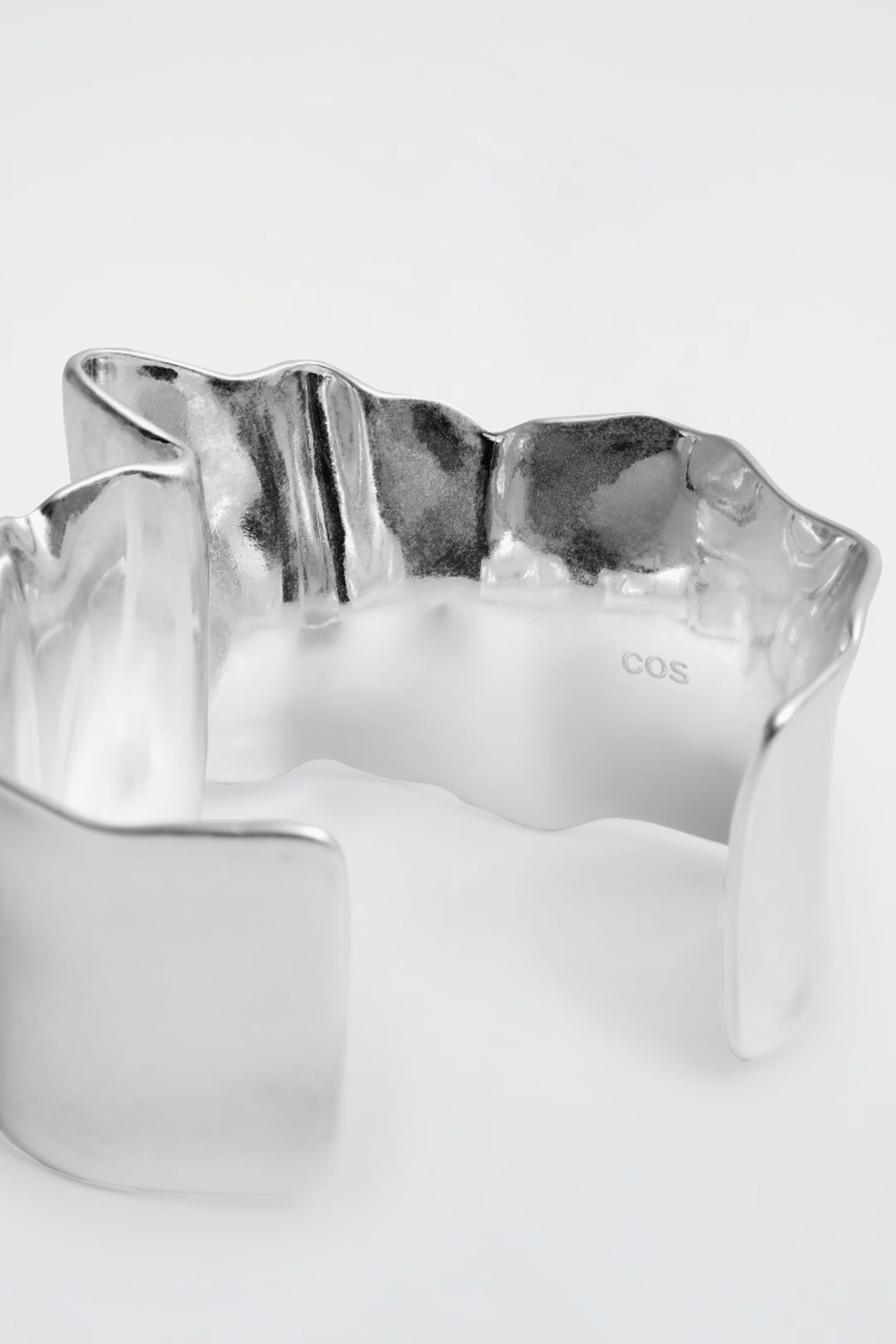 Curved silver cuff bracelet with a smooth, reflective surface and an engraved "COS" logo inside on a plain background.