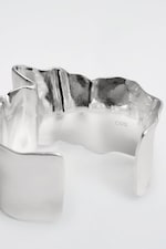 Placeholder for: Curved silver cuff bracelet with a smooth, reflective surface and an engraved "COS" logo inside on a plain background.