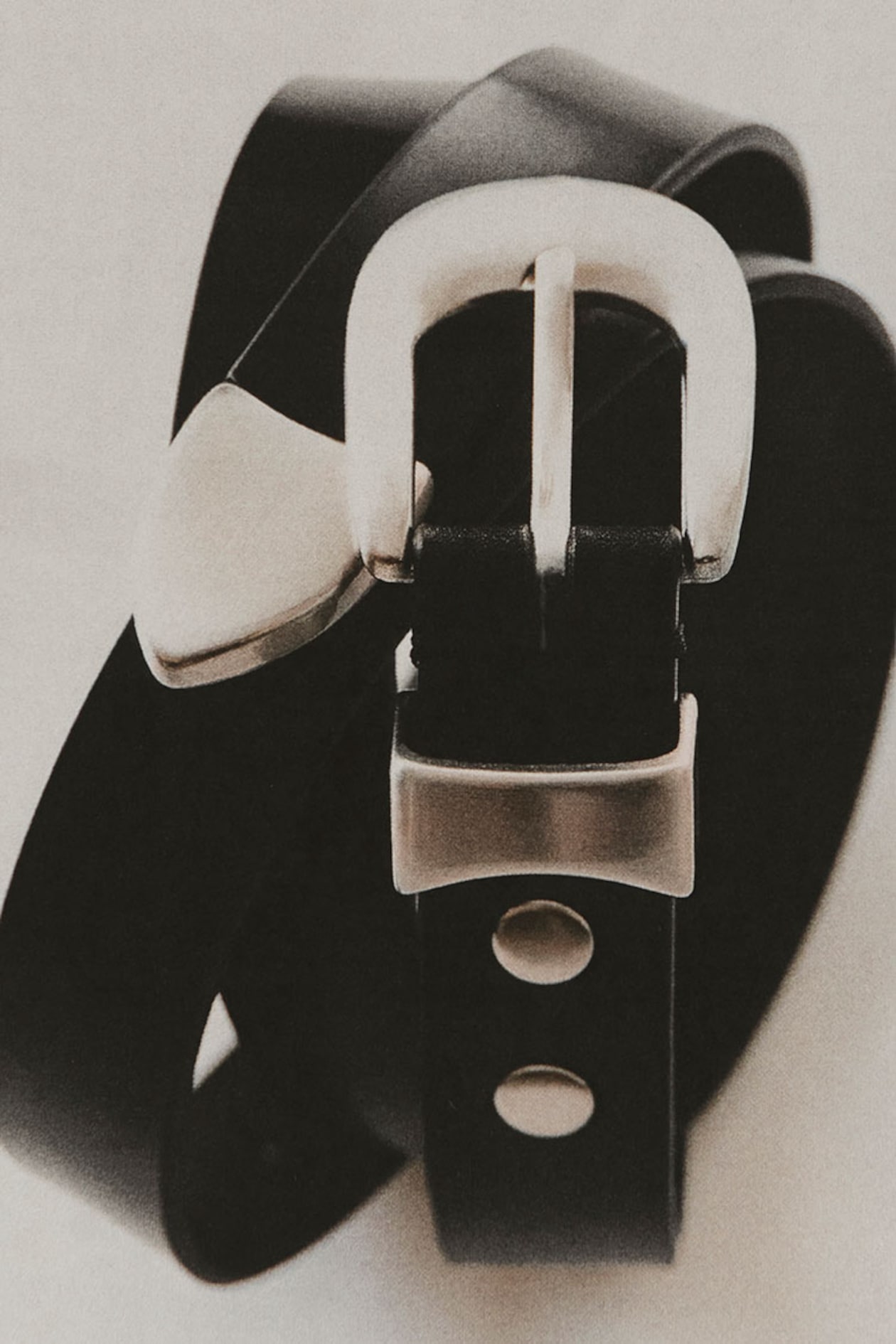 Close-up of a black leather belt with a silver buckle and rivet details, coiled in a spiral shape on a light background.