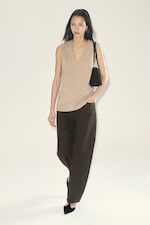 Placeholder for: Person in a beige sleeveless top, dark wide-leg pants, and black shoes, carrying a black shoulder bag, walking confidently on a light background.