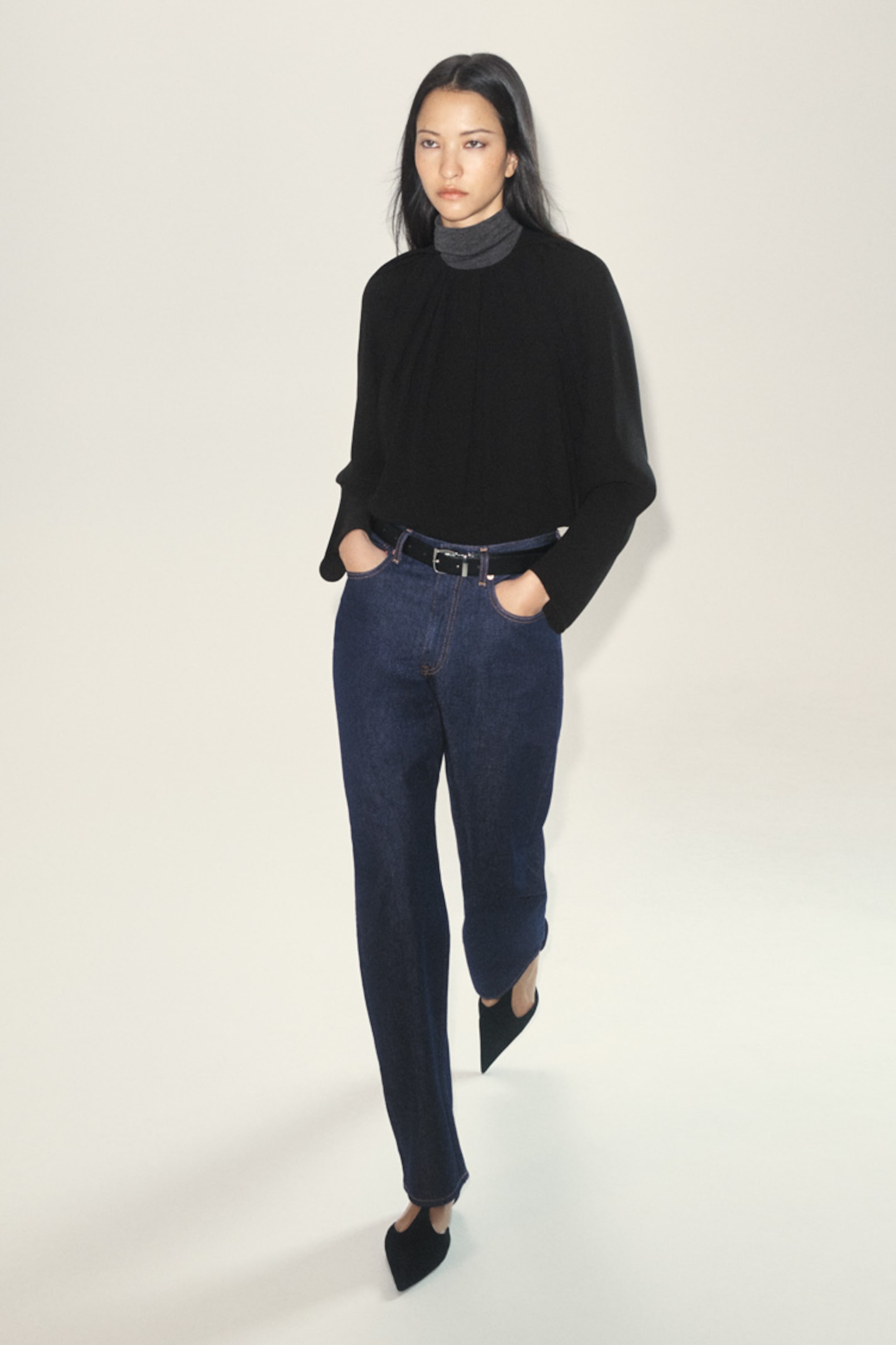 A woman in a black top and blue jeans walks confidently against a light background.