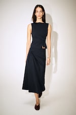 Placeholder for: Woman in a sleeveless, navy blue, asymmetric dress and brown shoes, standing against a plain white background.