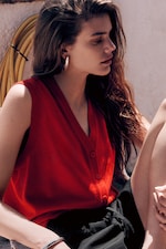 Placeholder for: Woman with long hair wearing a red sleeveless top and hoop earrings, sitting outdoors in sunlight with a yellow hose in the background.