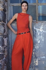 Placeholder for: Woman in a sleeveless red outfit with a black belt stands against a rustic metal door, wearing a black headband.