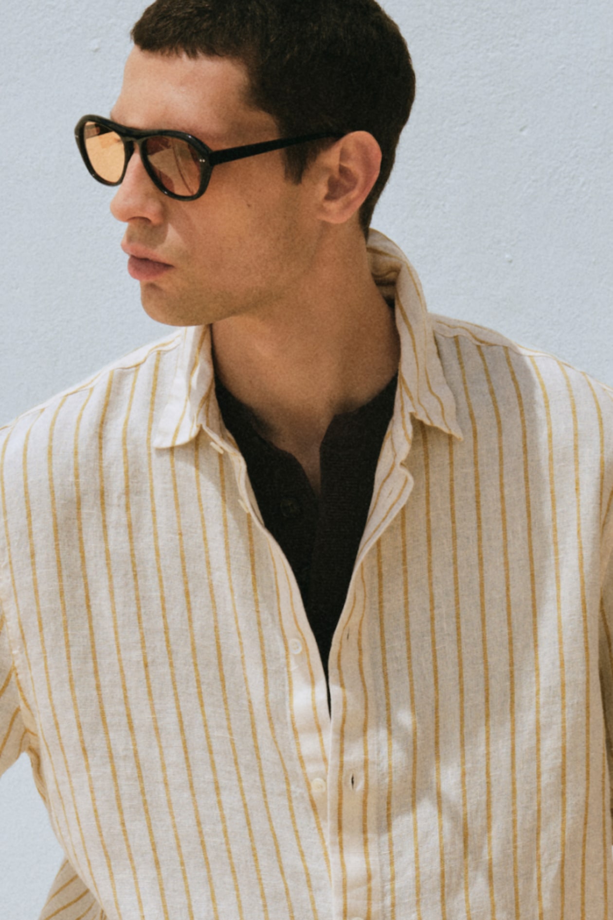 Man in sunglasses wearing a light-colored, striped button-up shirt, looking to the side against a light background.