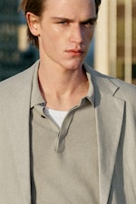 Placeholder for: Person in a light-colored blazer and polo, with short hair, gazing forward. The background is blurred, with vertical lines visible.