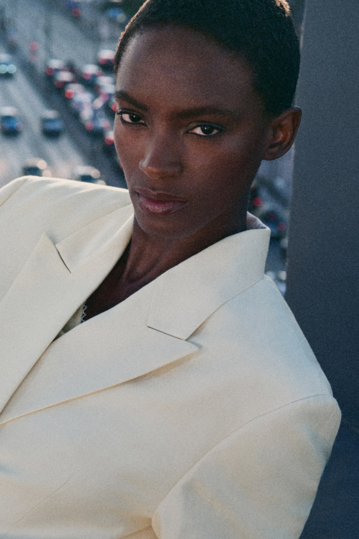 Person in a white suit with a serious expression, standing outdoors with blurred cityscape and traffic in the background.
