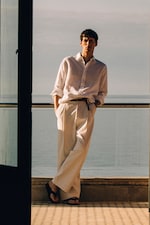 Placeholder for: Man in a white shirt and trousers leans against a railing, overlooking a calm sea under a clear sky.