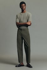 Placeholder for: Person in a grey shirt and olive trousers, arms crossed, standing against a plain background, wearing black shoes.