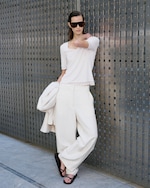 Placeholder for: WOMEN STANDING ON METAL FENCING, wearing white frilled t-shirt and white wide leg trouser