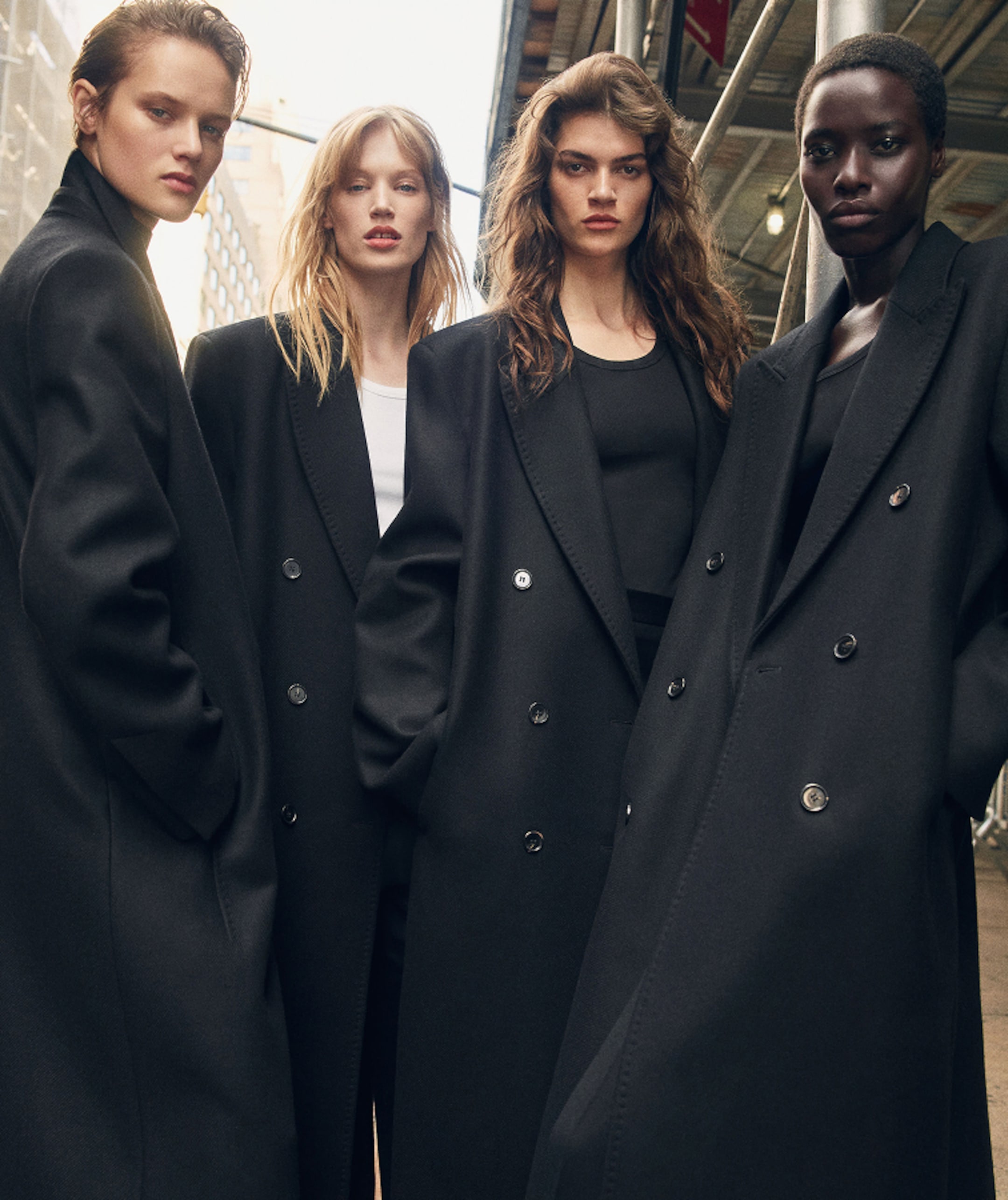 Four models wearing oversized black coats pose confidently outdoors, exuding a modern, stylish vibe under a partly cloudy sky.
