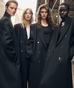 Placeholder for: Four models wearing oversized black coats pose confidently outdoors, exuding a modern, stylish vibe under a partly cloudy sky.