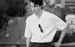 Placeholder for: Black and white photo of a person in a white button-up shirt with sunglasses hanging, gazing downward thoughtfully.