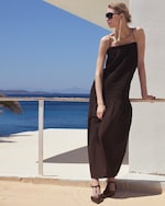 Placeholder for: Woman in a black dress and sunglasses, leaning against a white wall, with a sea view and blue sky in the background.