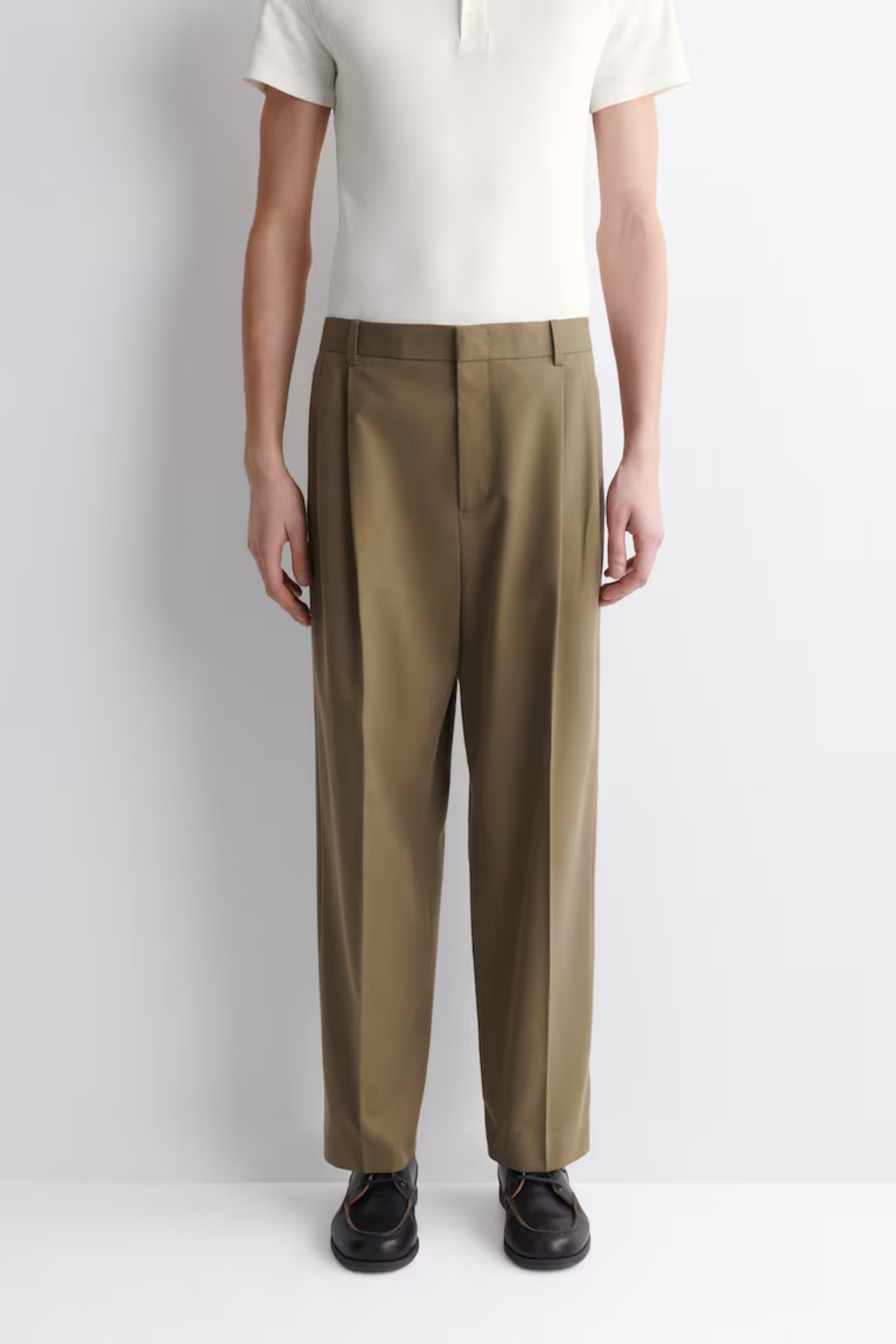 Person in a white polo shirt and olive green pleated trousers, wearing black shoes, standing against a plain light background.