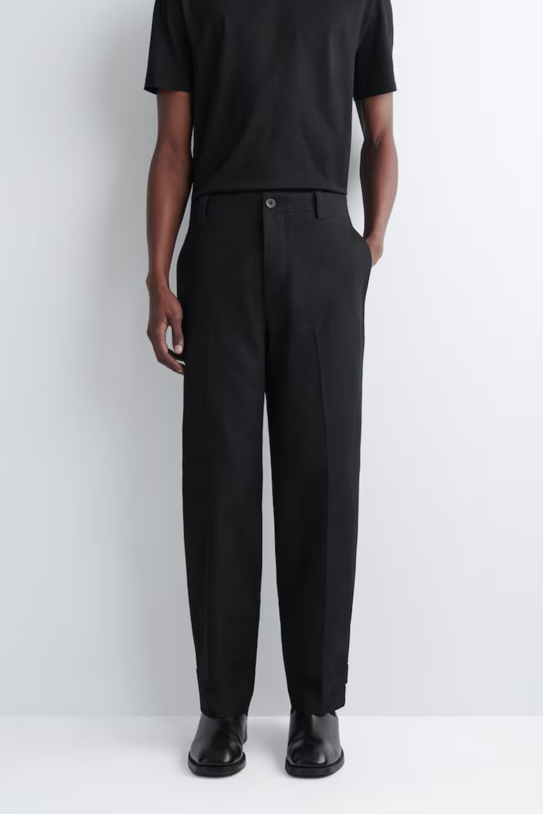 Man wearing black T-shirt, trousers, and shoes, standing against a white background with hands in pockets.