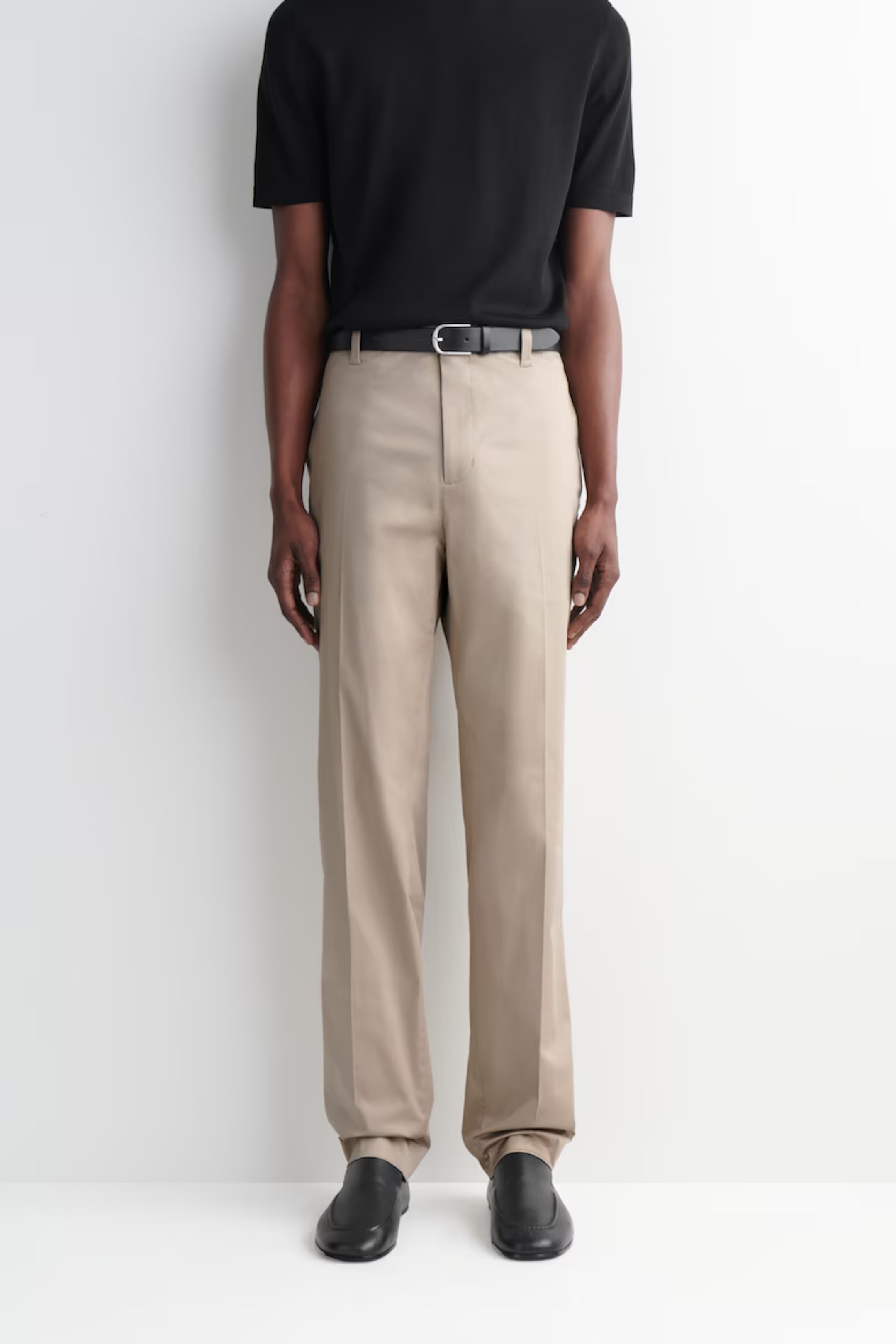 Man wearing a black T-shirt, beige trousers with a black belt, and black shoes, standing against a white background.