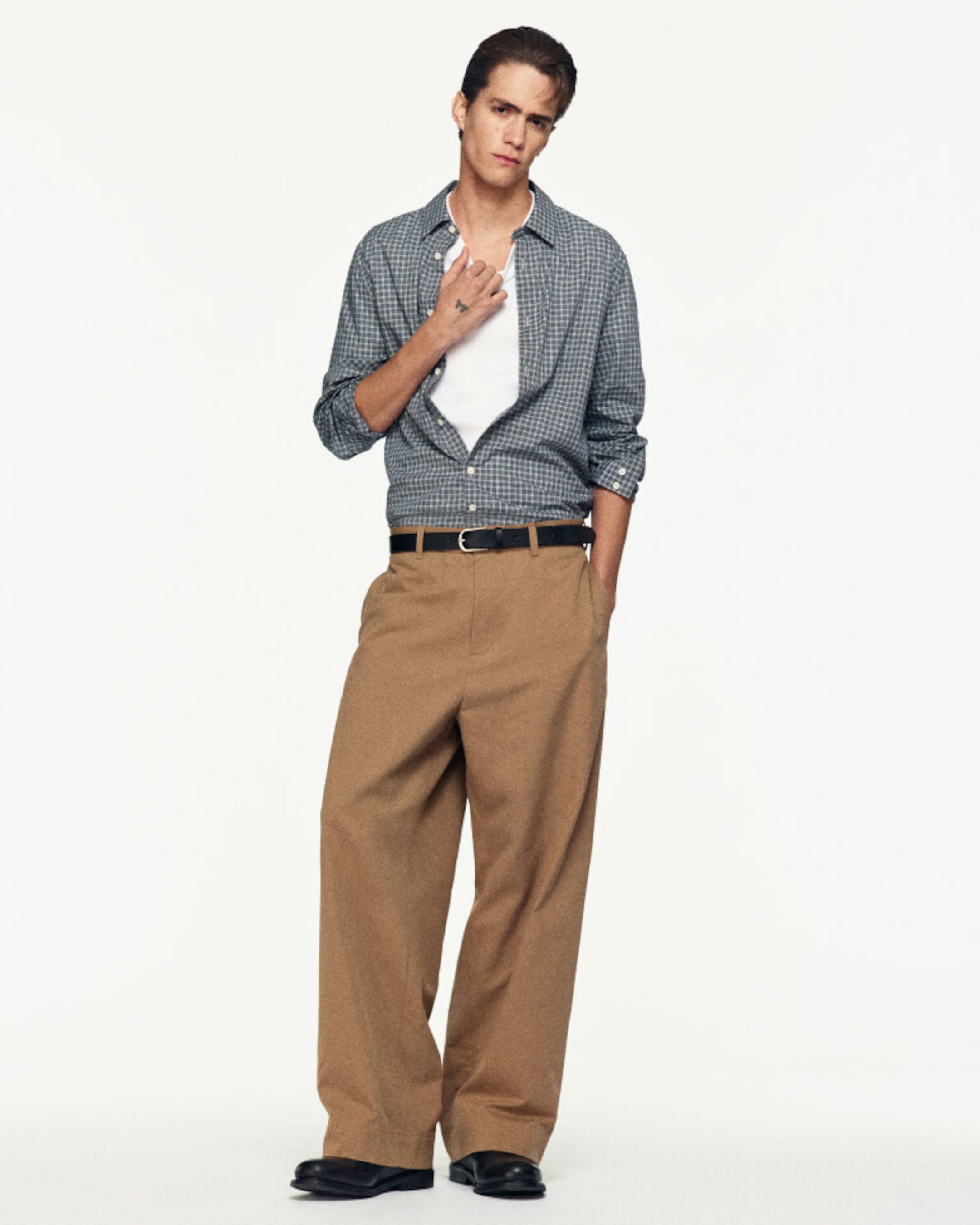 A person wearing a checkered shirt, white undershirt, brown wide-leg trousers, and black shoes poses confidently against a plain white background.